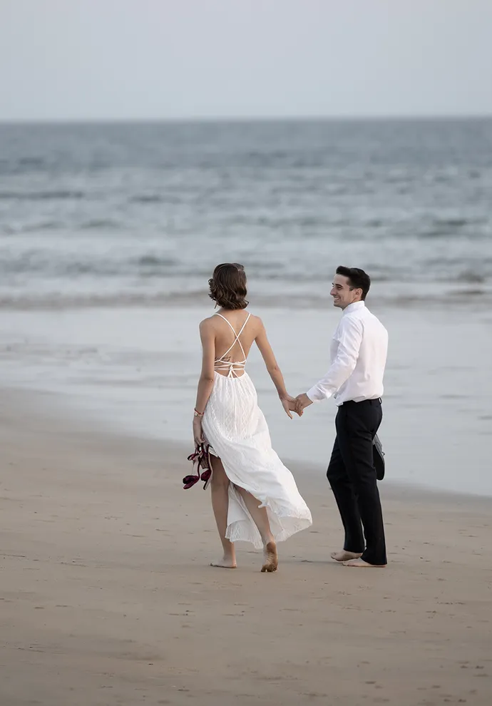 Estibaliz y Diego. Pareja de novios pasean de la mano en Playa Zahora. En sus manos llevan sus zapatos