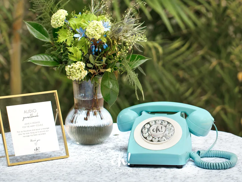 Teléfono audiolibro de firmas para invitados en una mesa con un cartel y unas flores Audio guestbook phone with flowers vase