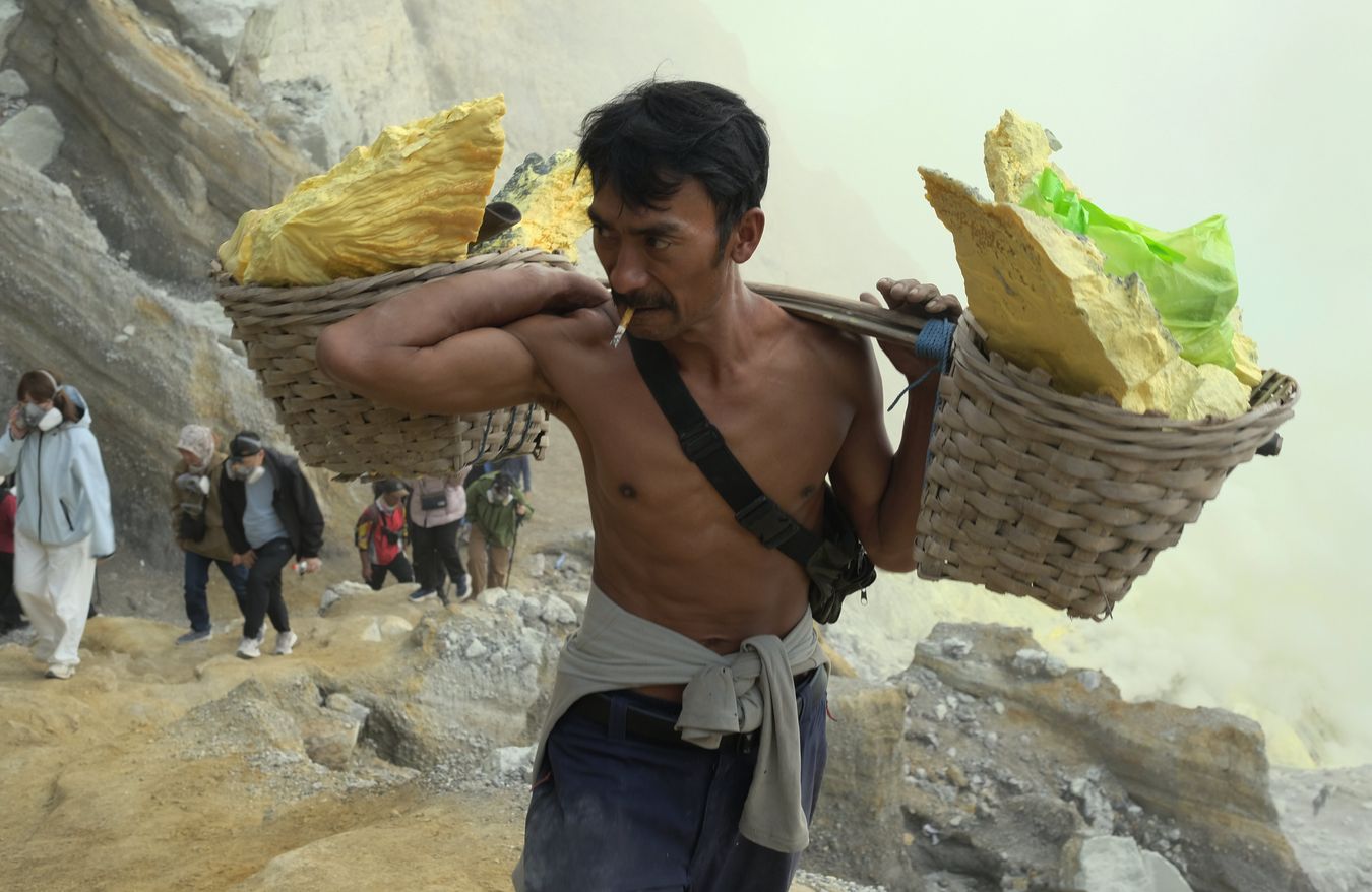 Miner ascending from the crater of Ijen volcano with traditional baskets loaded with sulfur, and followed by a group of tourists 